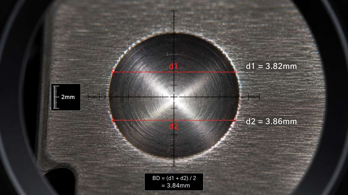 Close-up macro photograph of Brinell hardness test indentation on polished grey cast iron surface showing d1 and d2 diameter measurement per IS 1500 procedure.